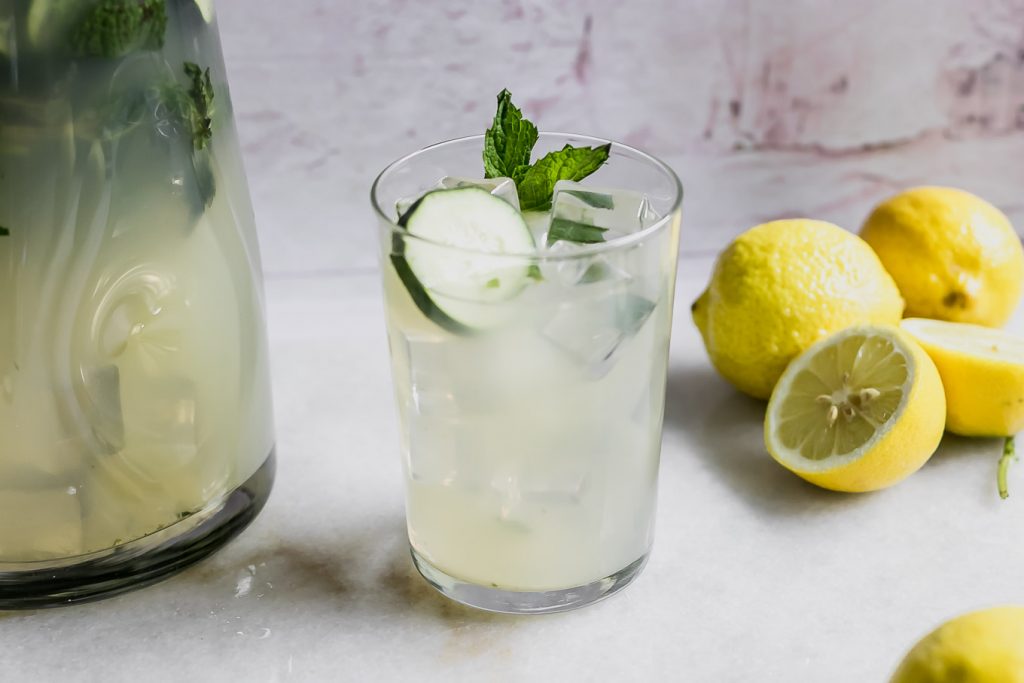 a glass of lemonade with cucumber and mint garnish on a table with lemons
