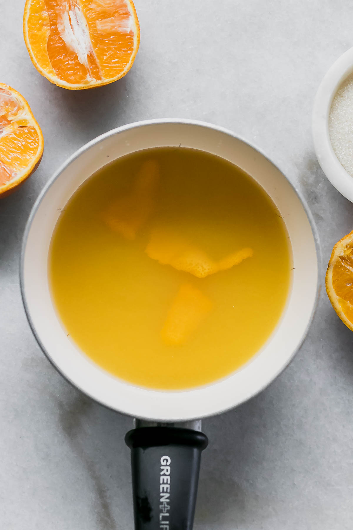 water, sugar, and orange juice in a saucepan on a white table