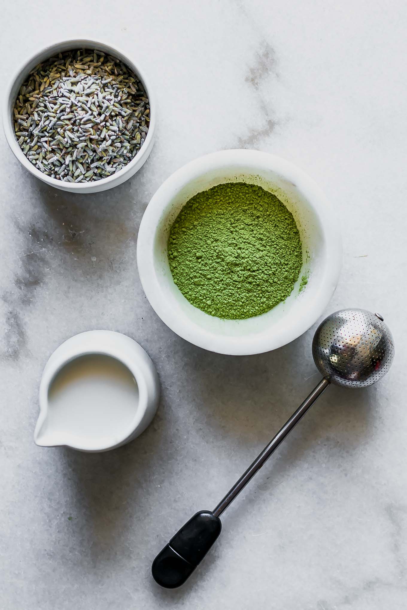 bowls of lavender, matcha green tea powder, and milk on a white table with a tea infuser