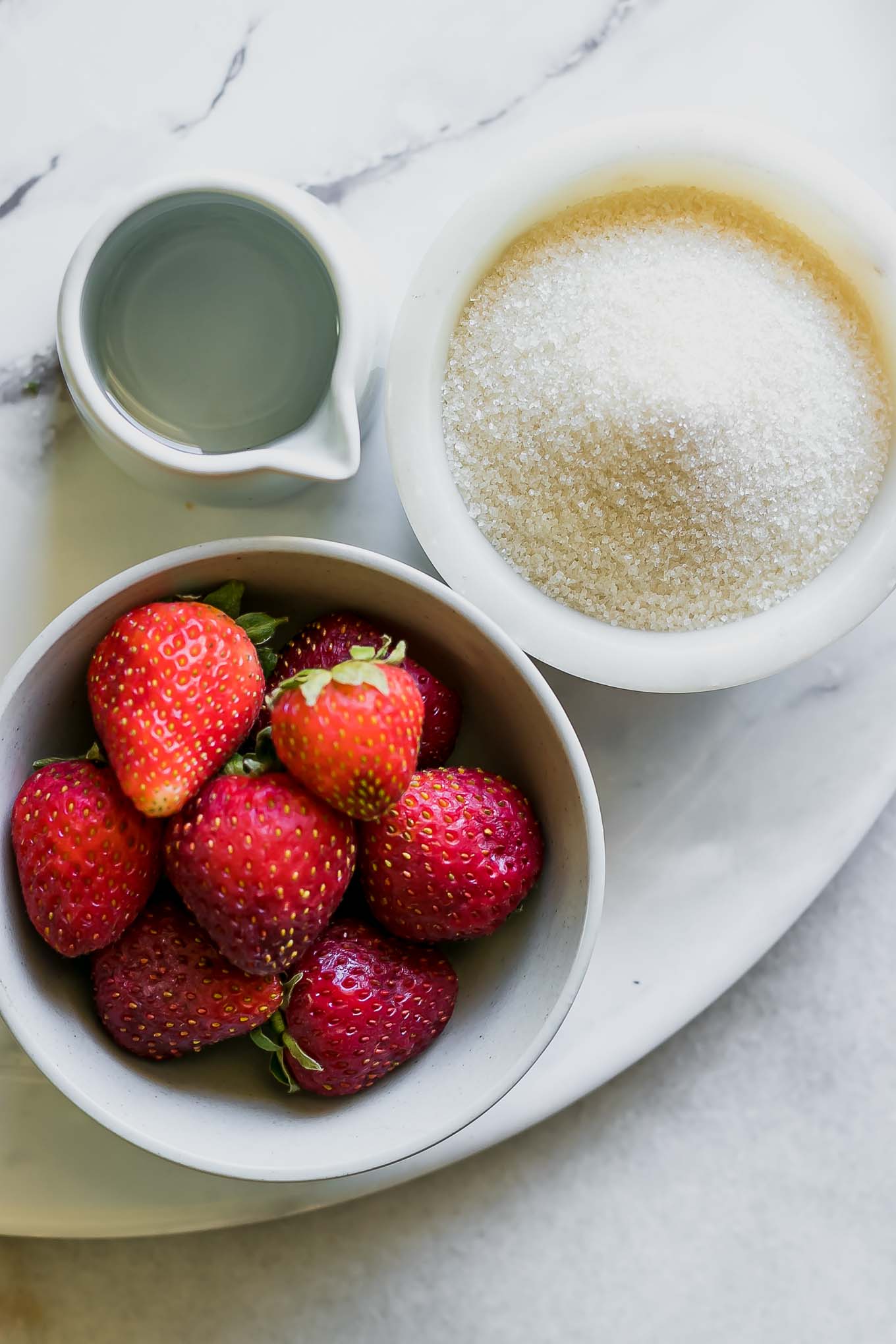 bowls of fresh strawberries, granulated sugar, and water on a white table for strawberry syrup